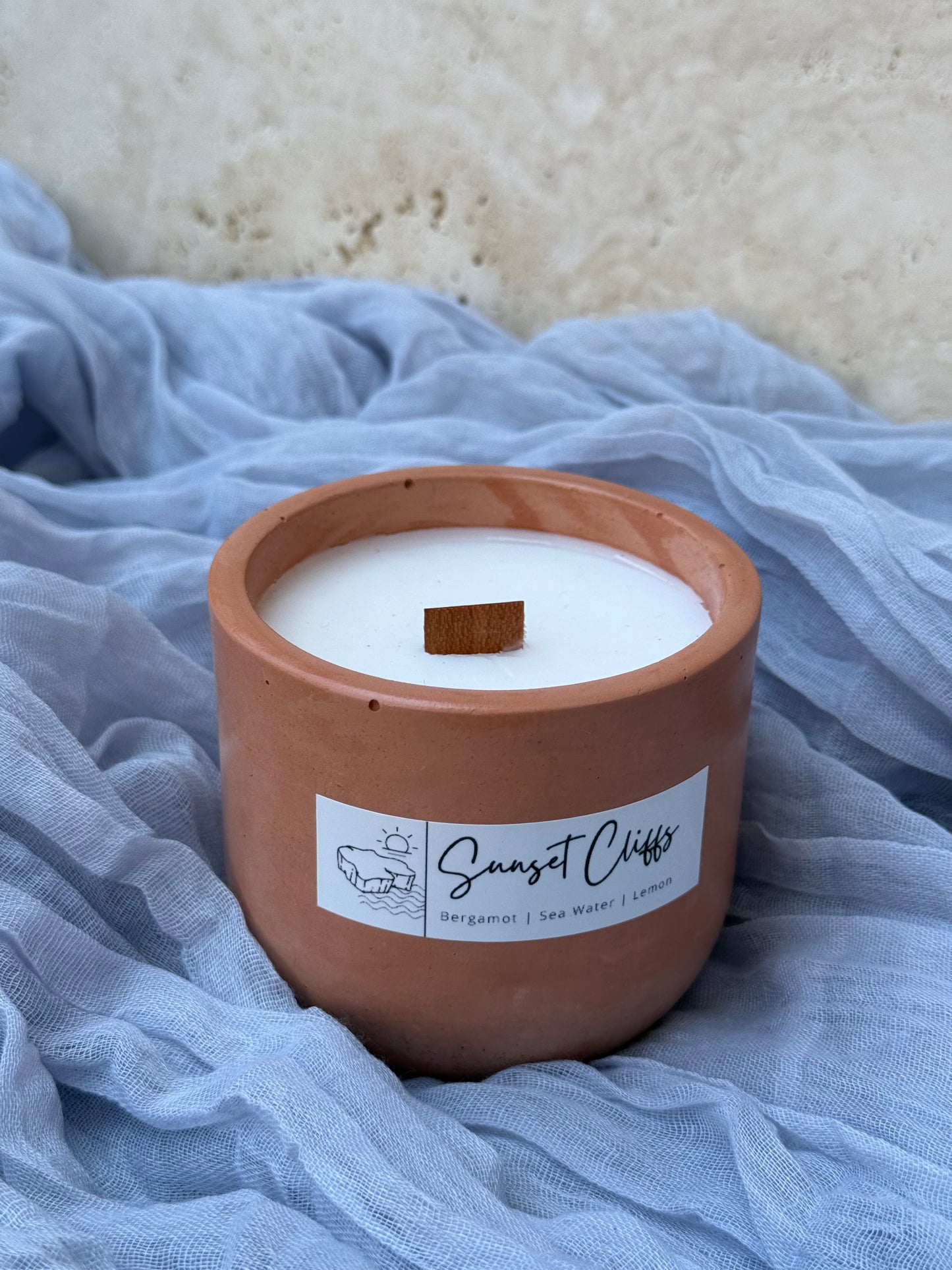 Candle in a terracotta pot labeled 'Sunset Cliffs' on a textured blue fabric background.